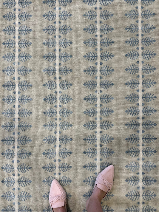 A person's feet wearing light pink shoes standing on a blue, beige and gray patterned modern oushak hand- knotted wool rug.