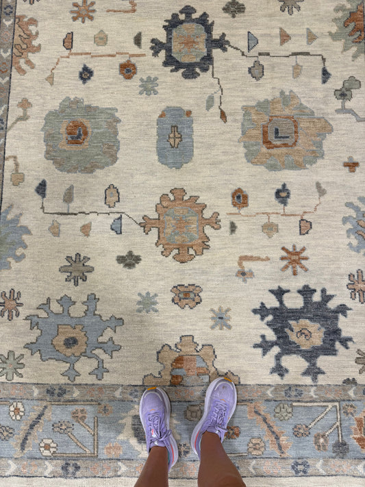 Neutral background with light blue border turkish oushak rug with accents of navy, coral, orange, light blue, and beige.