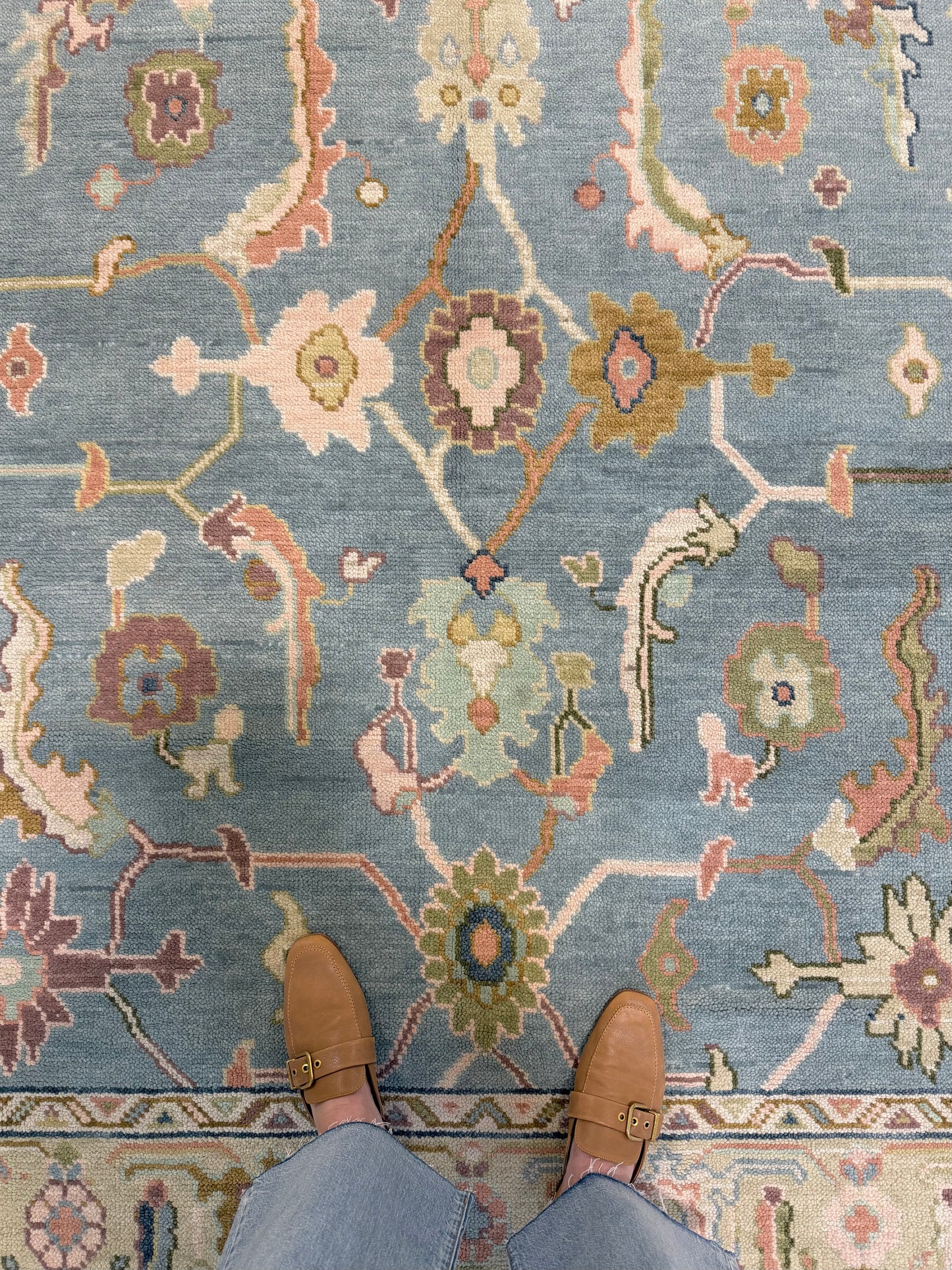 A top-down view of a patterned hand-knotted Oushak wool rug with a blue foundation and multiple colors including beige, brown, and green, with a person's feet wearing brown shoes visible at the bottom of the frame.