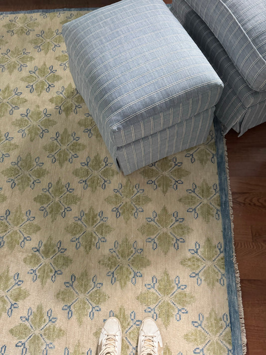 A modern Oushak area rug with a warm gray background and pops of color from cobalt blue, chartreuse, and light coral, displayed in a room setting with a person's feet and a blue cushion for scale.