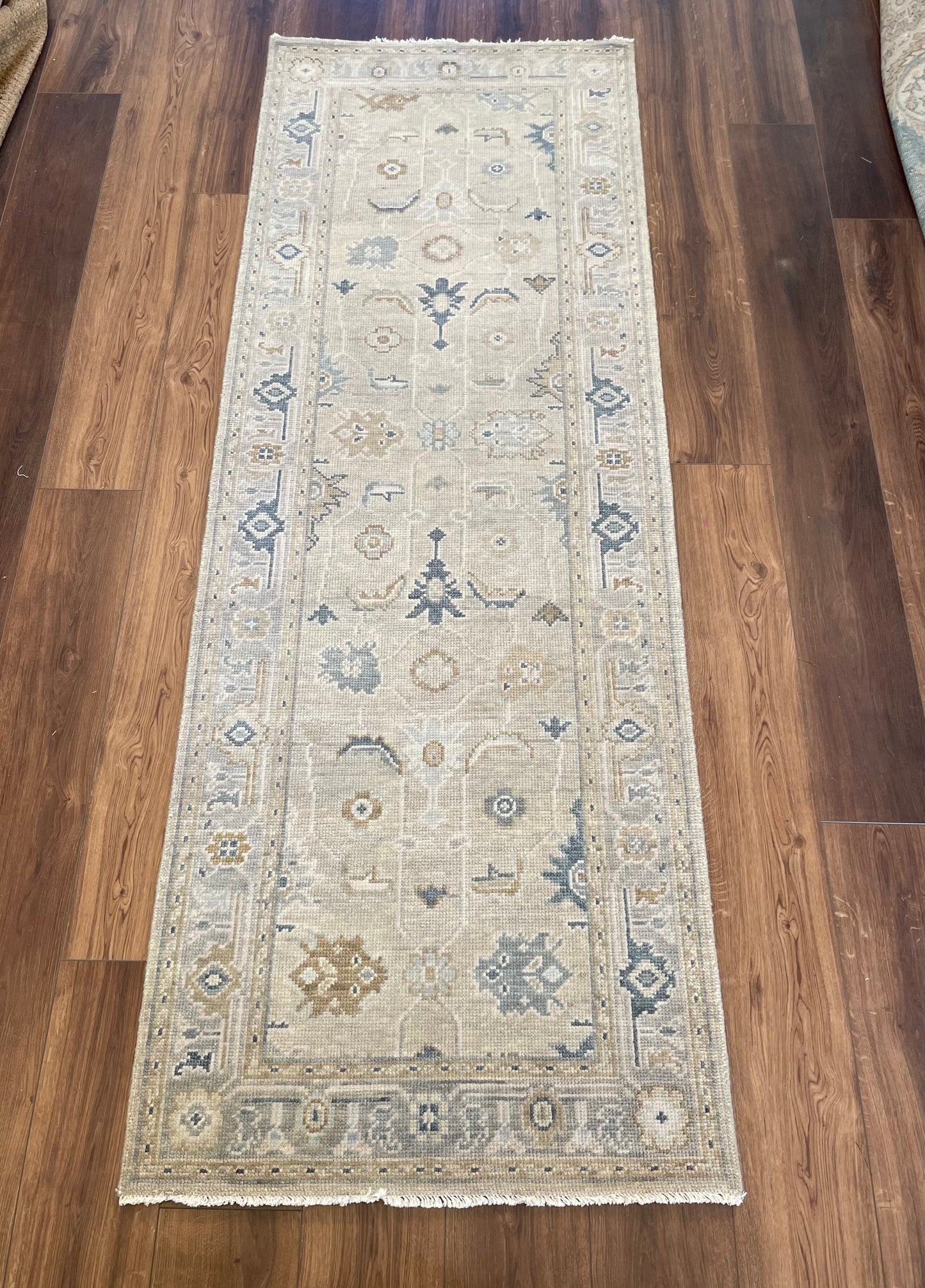 A person standing on a modern Oushak rug with a soft beige base featuring geometric patterns in gray, cream, and blue. Only the person's feet and part of their legs are visible.