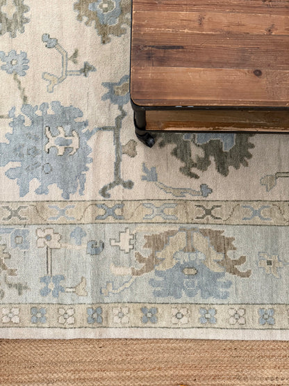 Evangeline warm blue and green oushak rug layered with jute rug and wood table atop