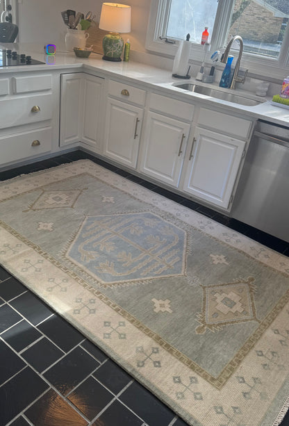 Lennon Blue and green 100% wool Modern Oushak rug with neutral border and green accents and blue center medallion in kitchen with white cabinets.