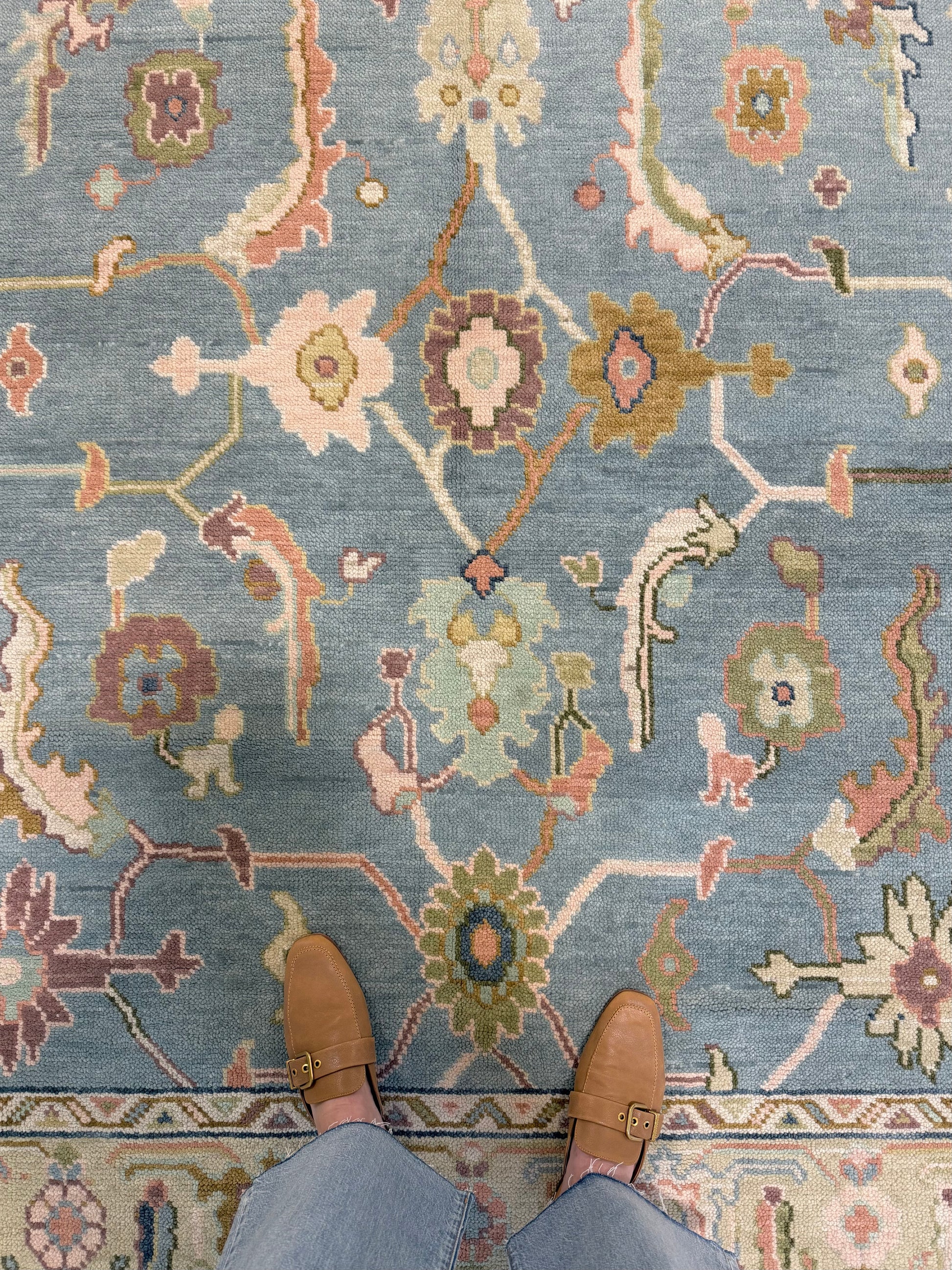 A top-down view of a patterned hand-knotted Oushak wool rug with a blue foundation and multiple colors including beige, brown, and green, with a person's feet wearing brown shoes visible at the bottom of the frame.