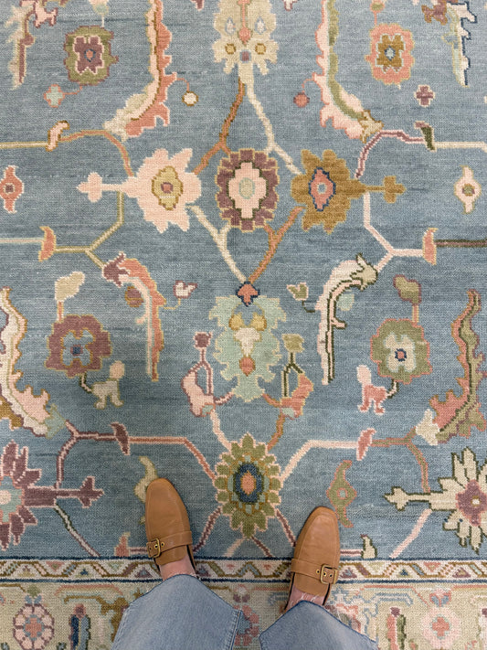 A top-down view of a patterned hand-knotted Oushak wool rug with a blue foundation and multiple colors including beige, brown, and green, with a person's feet wearing brown shoes visible at the bottom of the frame.