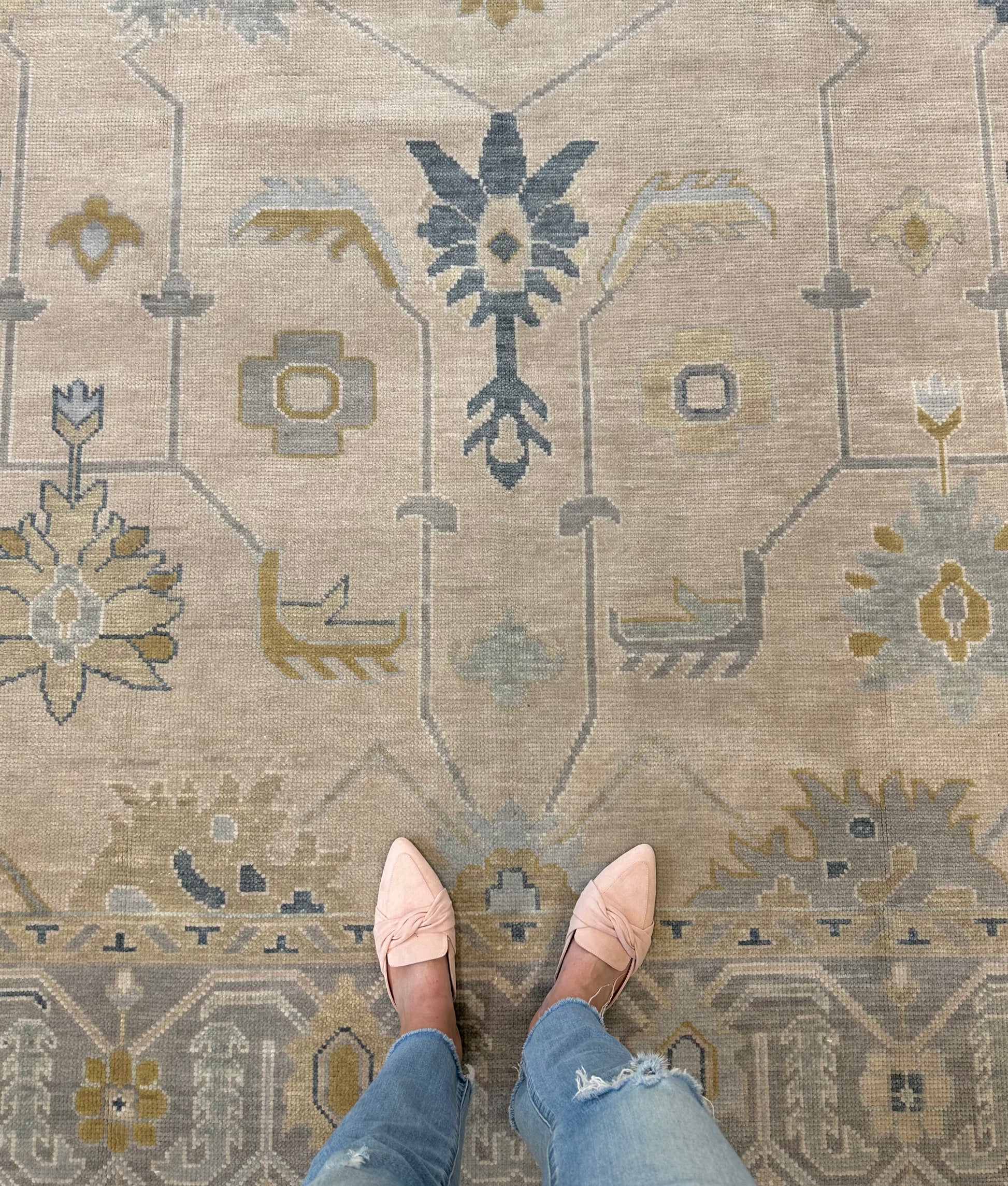 A person's feet wearing light pink shoes and blue jeans, standing on a traditional style beige, gray, blue, and green Oushak rug.