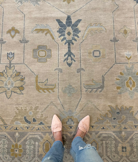 A person's feet wearing light pink shoes and blue jeans, standing on a traditional style beige, gray, blue, and green Oushak rug.
