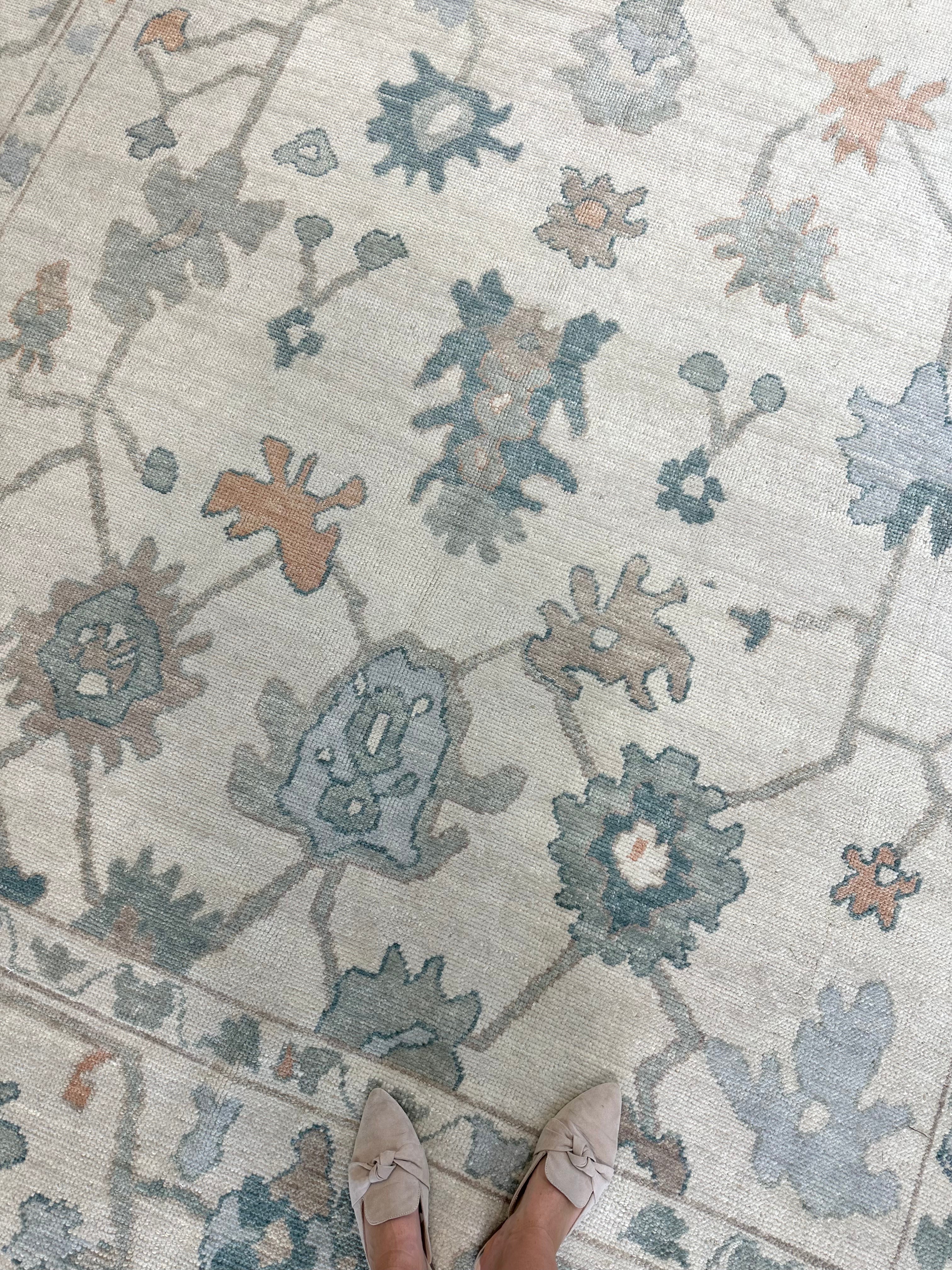 A close-up view of Olivia Turkish Oushak rug with a floral pattern in off-white, gray, teal, orange, and light blue colors, with a person's feet in light-colored shoes visible at the bottom of the frame.