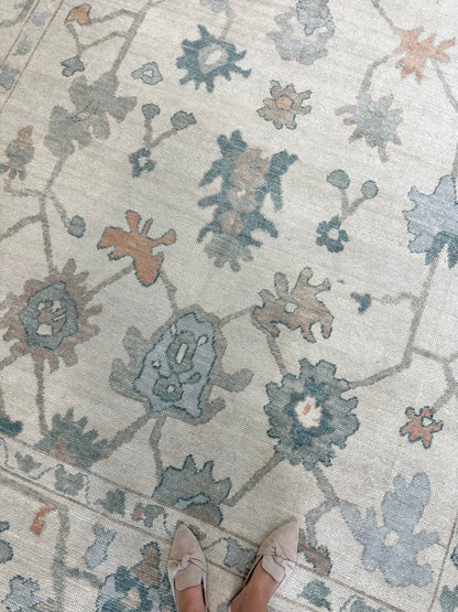 A close-up view of Olivia Turkish Oushak rug with a floral pattern in off-white, gray, teal, orange, and light blue colors, with a person's feet in light-colored shoes visible at the bottom of the frame.