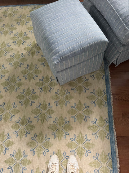 A modern Oushak area rug with a warm gray background and pops of color from cobalt blue, chartreuse, and light coral, displayed in a room setting with a person's feet and a blue cushion for scale.