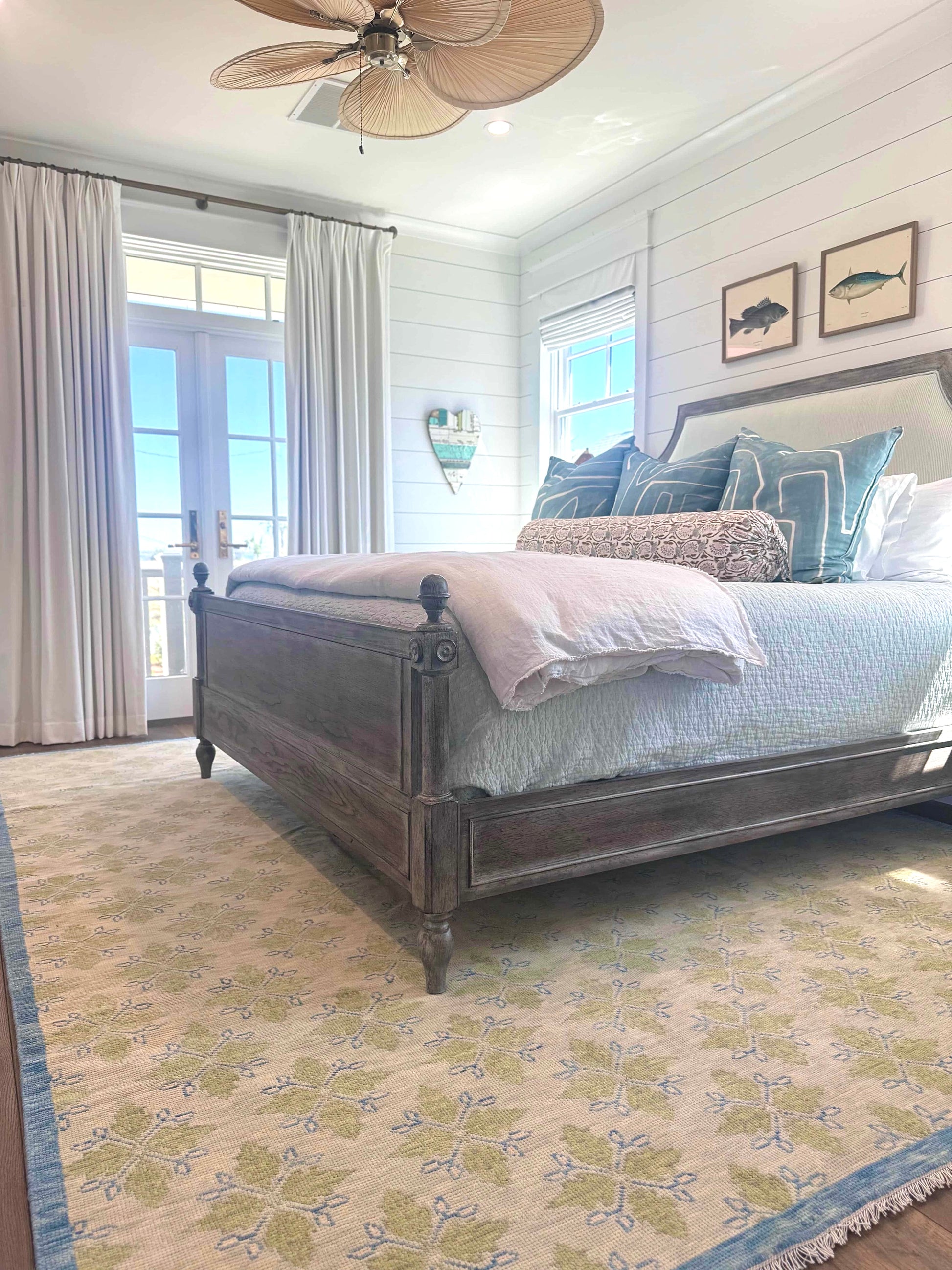A modern Oushak area rug with a warm gray background and pops of color from cobalt blue, chartreuse, and light coral, displayed in a coastal bedroom setting with a wooden bed and fish art above.