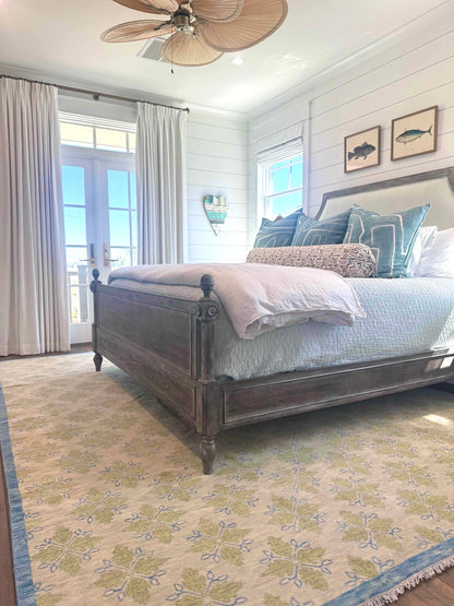 A modern Oushak area rug with a warm gray background and pops of color from cobalt blue, chartreuse, and light coral, displayed in a coastal bedroom setting with a wooden bed and fish art above.
