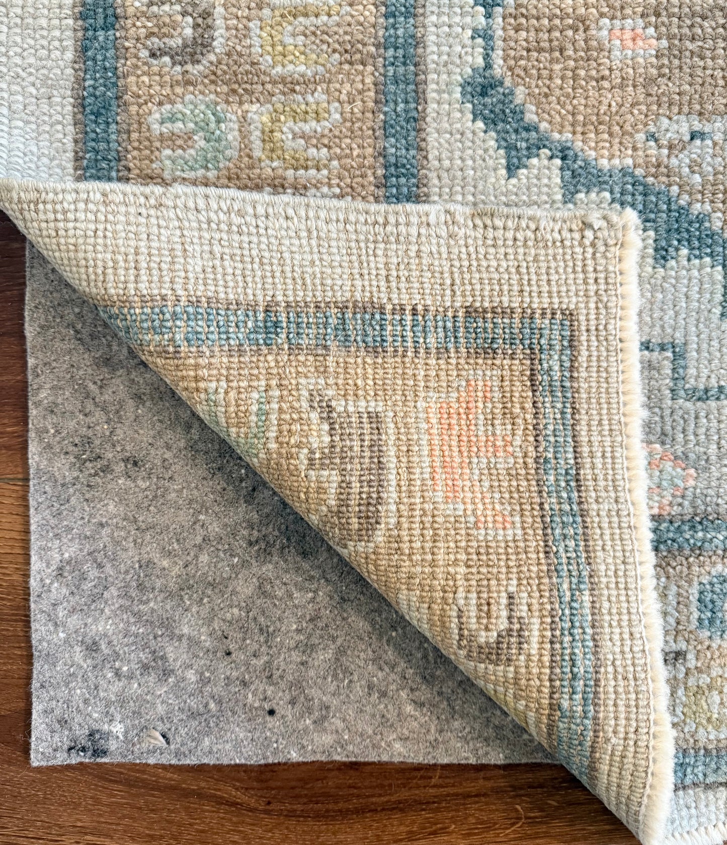 Close-up of a patterned hand-knotted turkish oushak  rug being rolled out on a wooden floor with rug pad underneath.