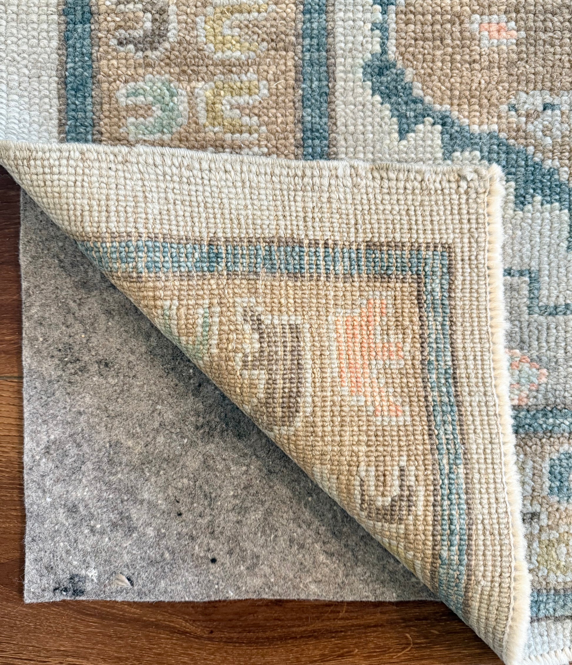 Close-up of a patterned hand-knotted turkish oushak  rug being rolled out on a wooden floor with rug pad underneath.