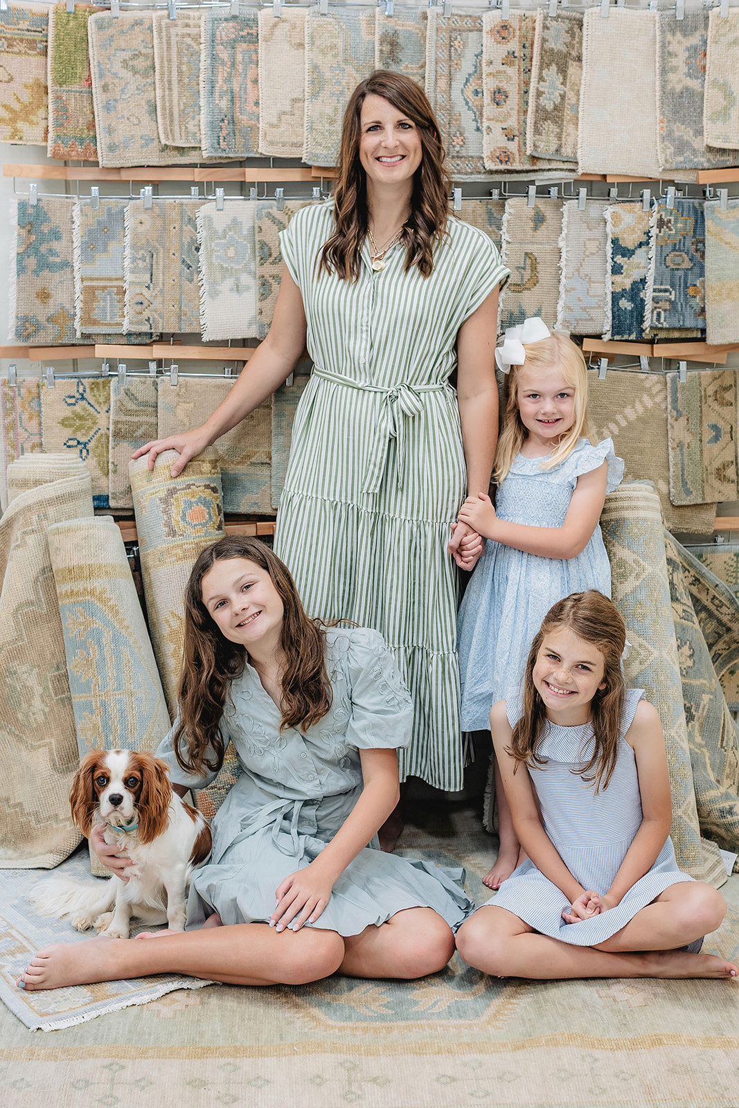 Woman and three young girls with a dog sitting on a rug in front of a wall of rugs.