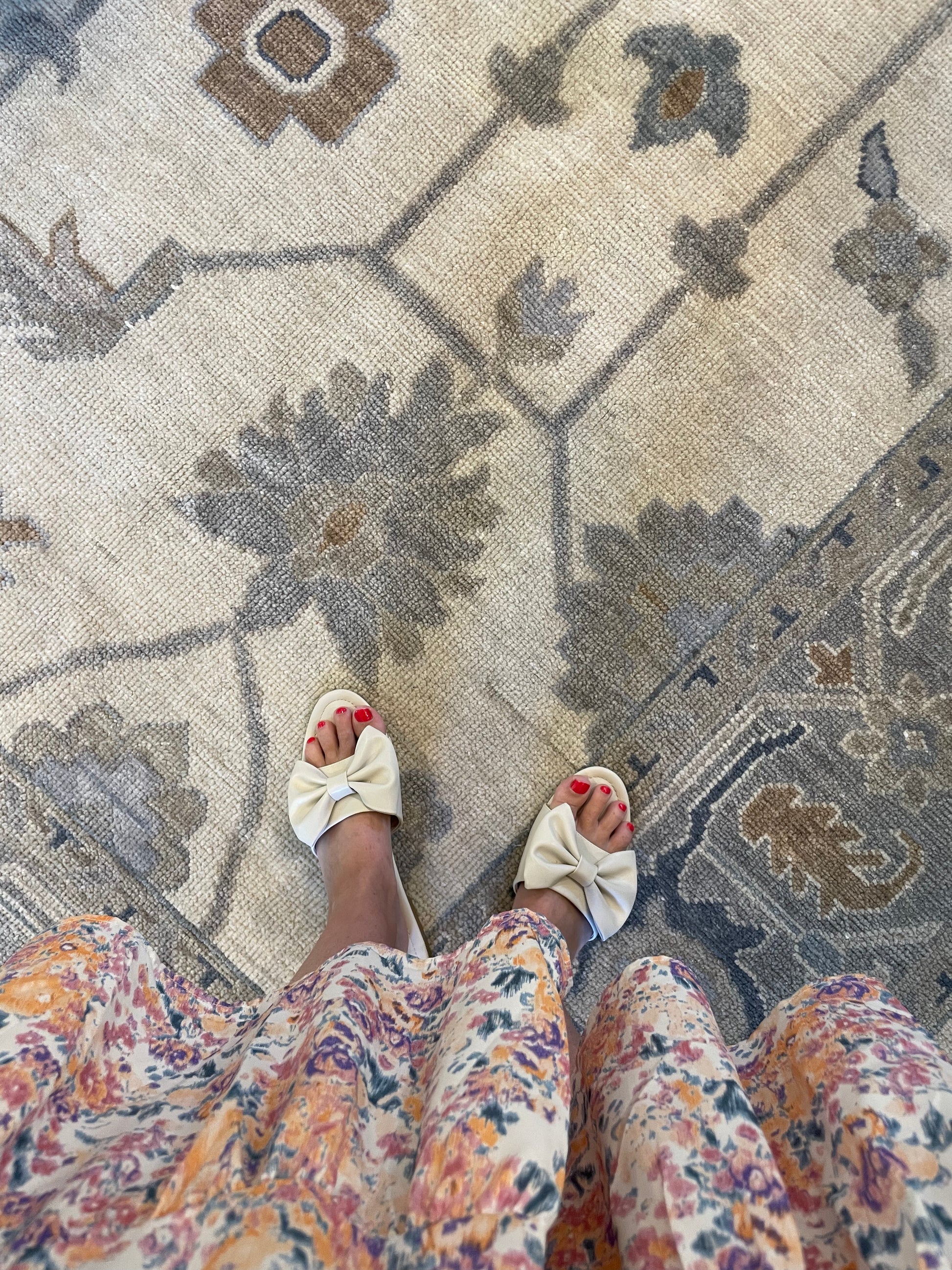 A view of a person's feet wearing floral patterned shoes, standing on a traditional style multicolored area rug with a floral pattern.