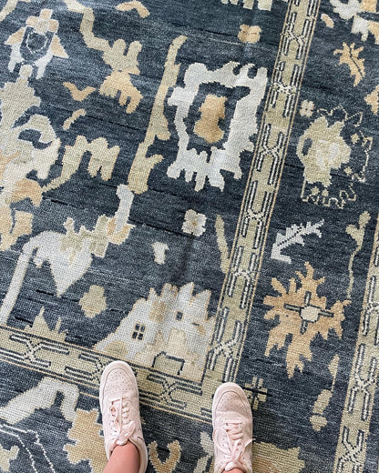 A person standing on a Eloise Modern  Oushak rug with a moody blue-green foundation, featuring accents of cream, olive, light blue, and tan.