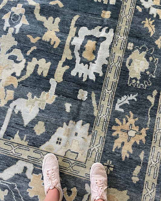 A person standing on a Eloise Modern  Oushak rug with a moody blue-green foundation, featuring accents of cream, olive, light blue, and tan.